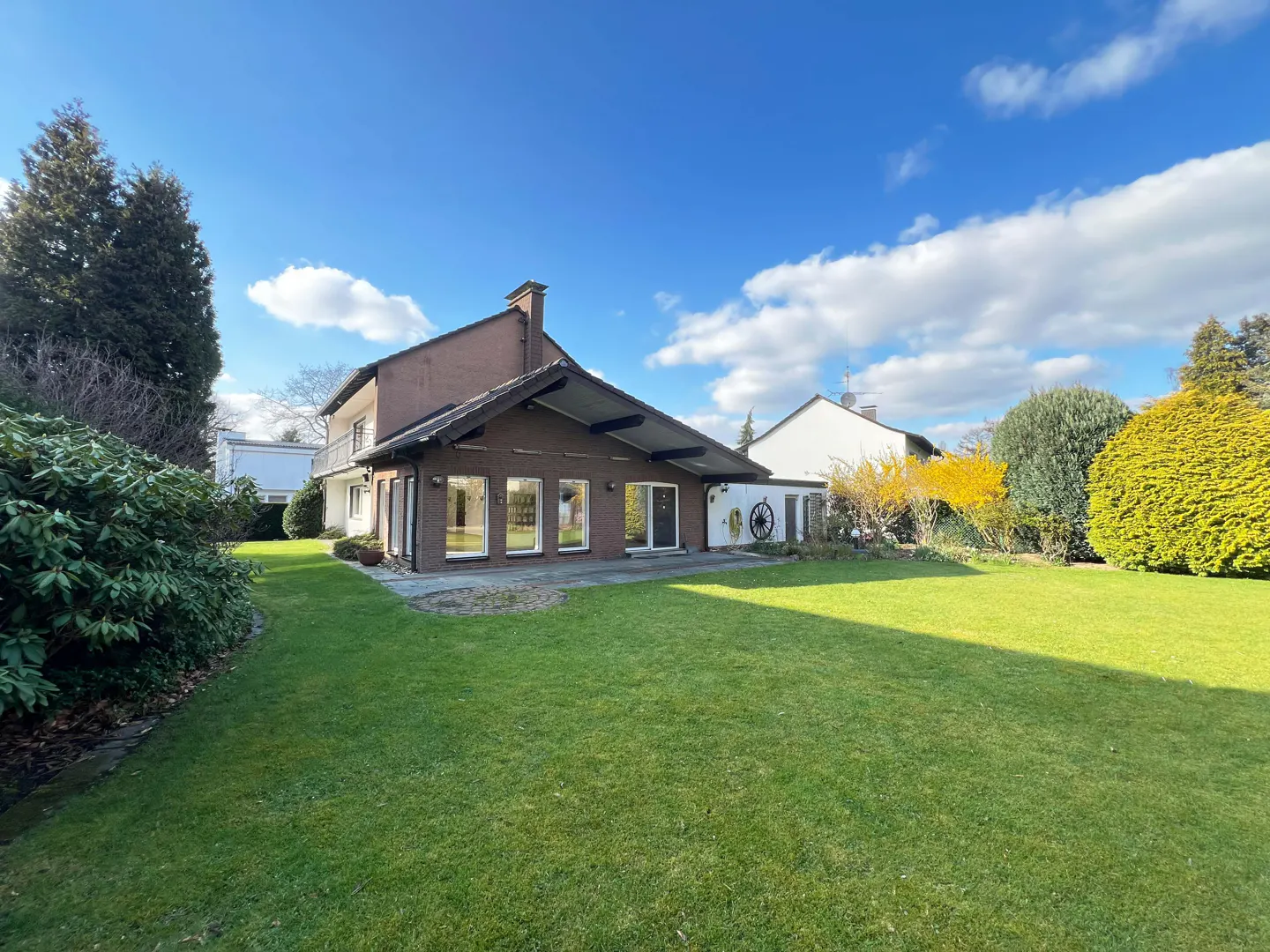 Backyard view of a brown two-story house with a large green lawn under a blue sky with white clouds.