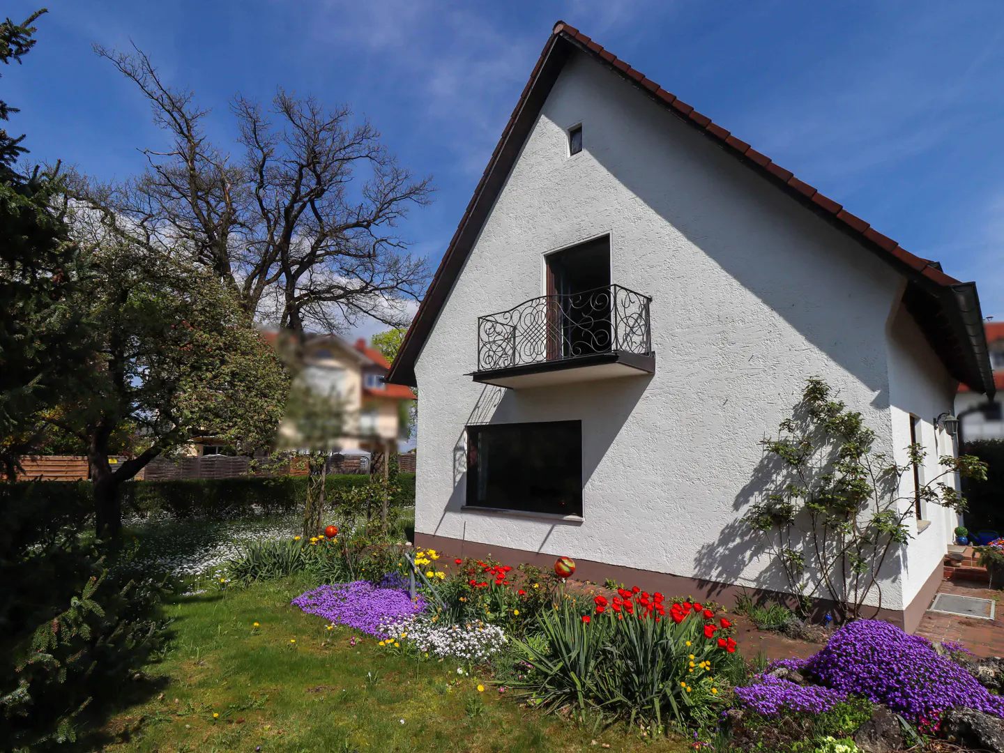 A white house with a brown roof and a small balcony, surrounded by a garden with colorful flowers.