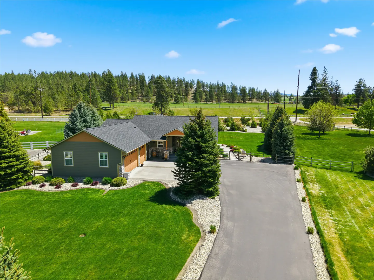 Aerial view of a green house with a gray roof, a long driveway, and a large green lawn. Trees and a blue sky are in the background.
