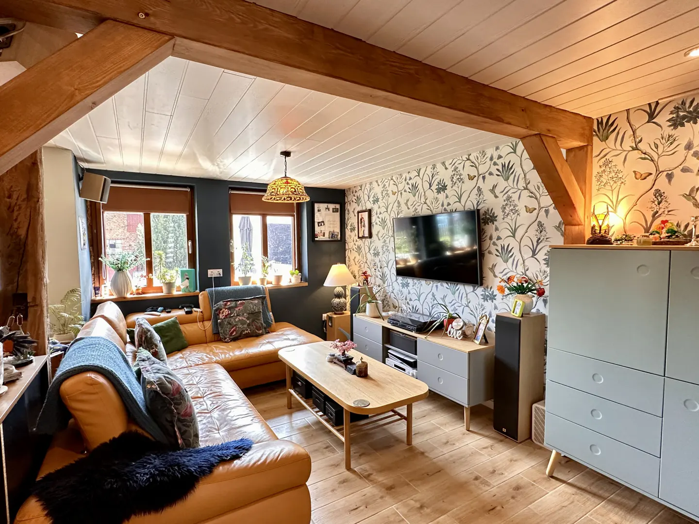 Cozy living room with exposed beams, floral wallpaper, and a tan leather sectional sofa. A flat-screen TV hangs above a gray media console.