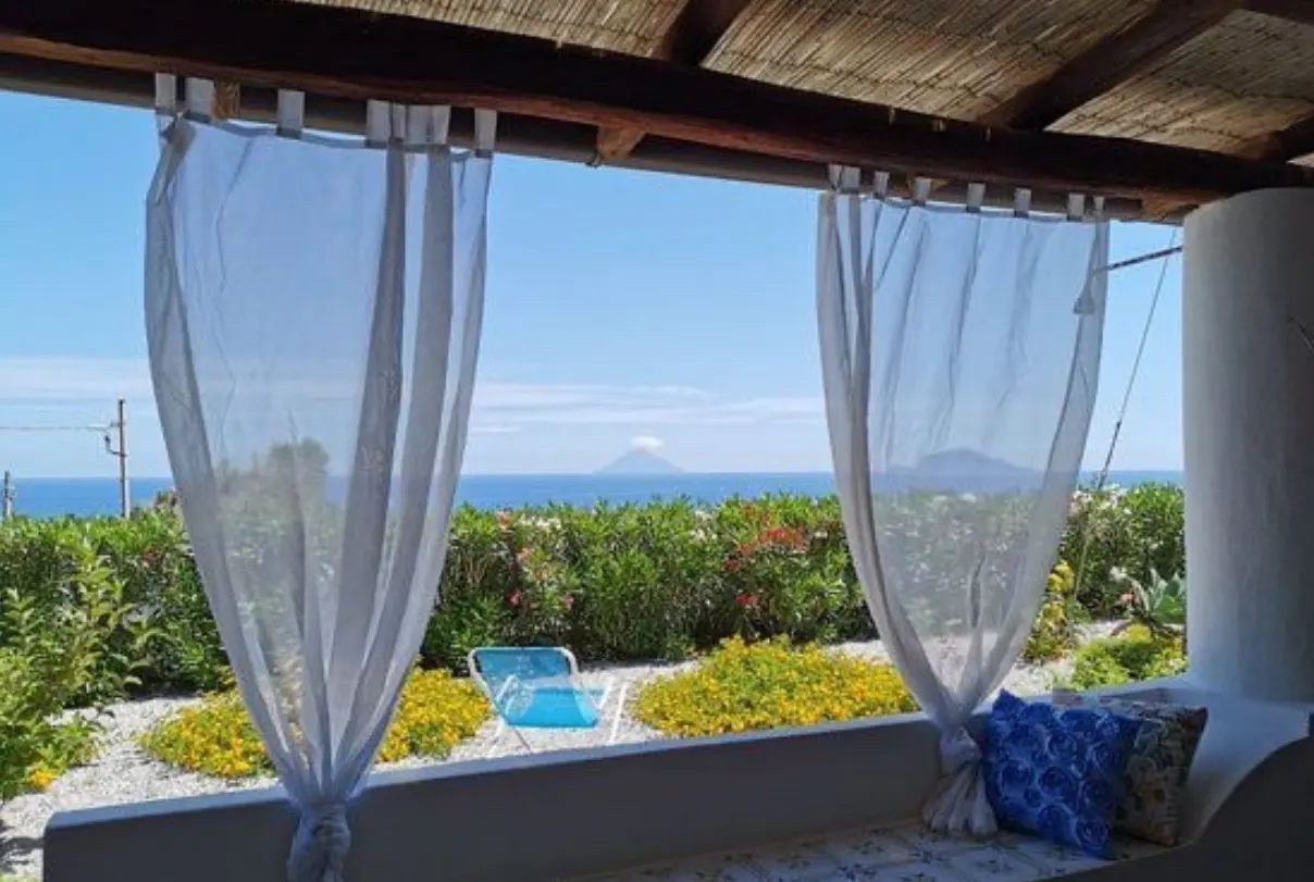 View from a shaded patio with white curtains, looking out over a garden, blue sea, and a distant volcanic island.