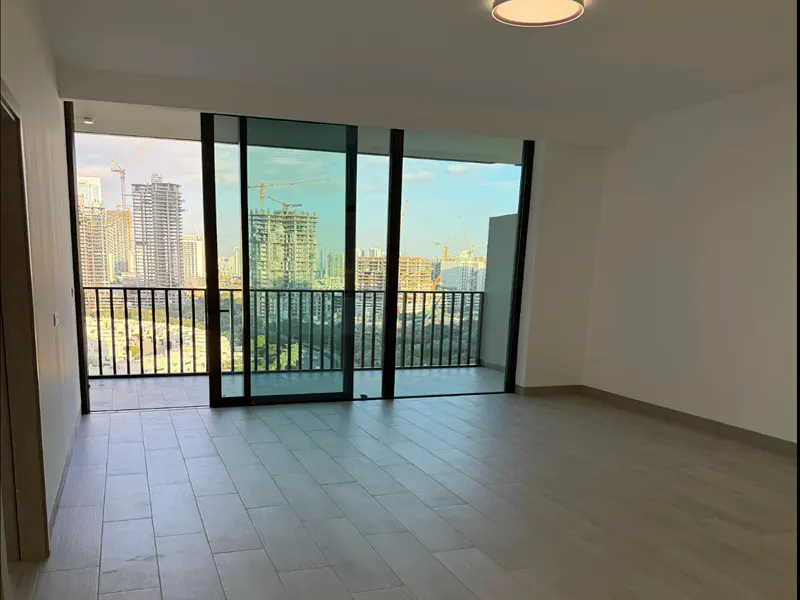 An empty room with light wood floors and white walls. A sliding glass door leads to a balcony with a city view.