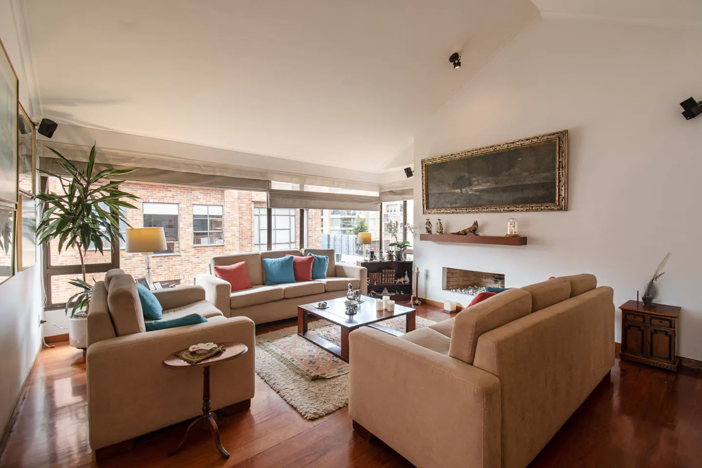 Living room with hardwood floors, beige sofas with blue and red pillows, and a fireplace with a painting above it.