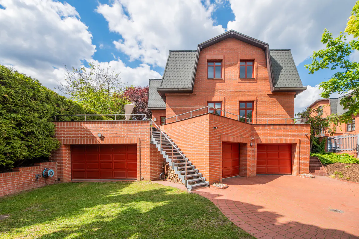 Three-story red brick house with three garage doors, an outdoor staircase, and a paved driveway under a blue sky.