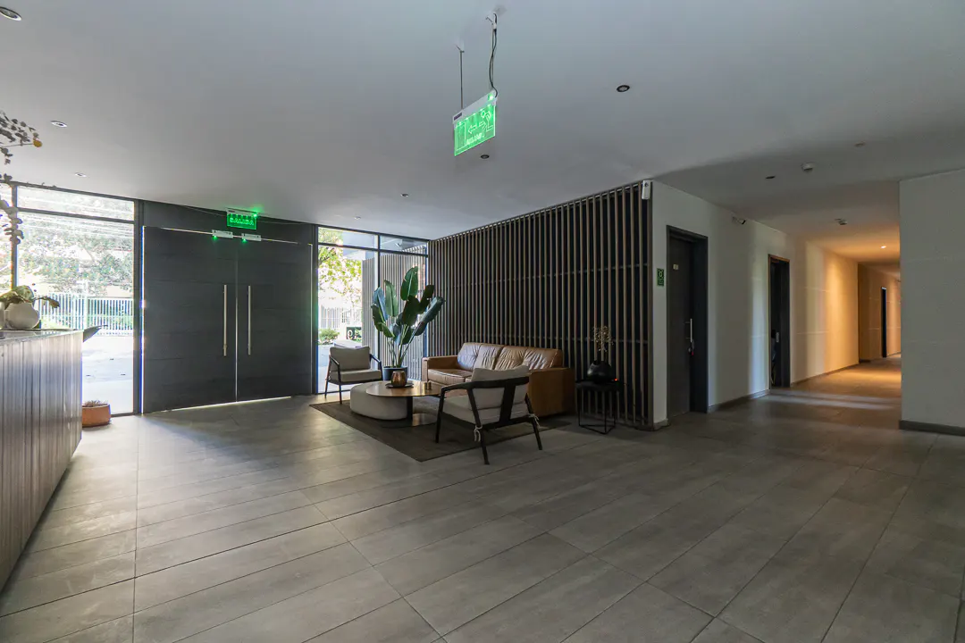 Modern lobby with gray tile floors, a brown leather sofa, and two chairs. A large plant sits near a window. An exit sign hangs from the ceiling.
