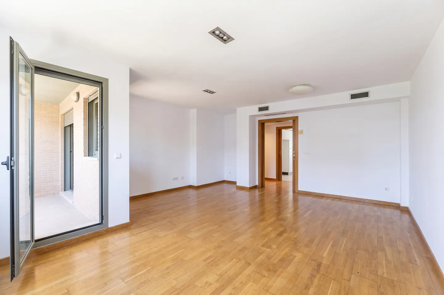 Bright, empty room with hardwood floors and white walls. An open glass door leads to a balcony.