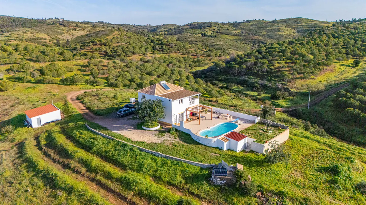 Aerial view of a white house with a pool, surrounded by green hills and trees. Cars parked nearby.