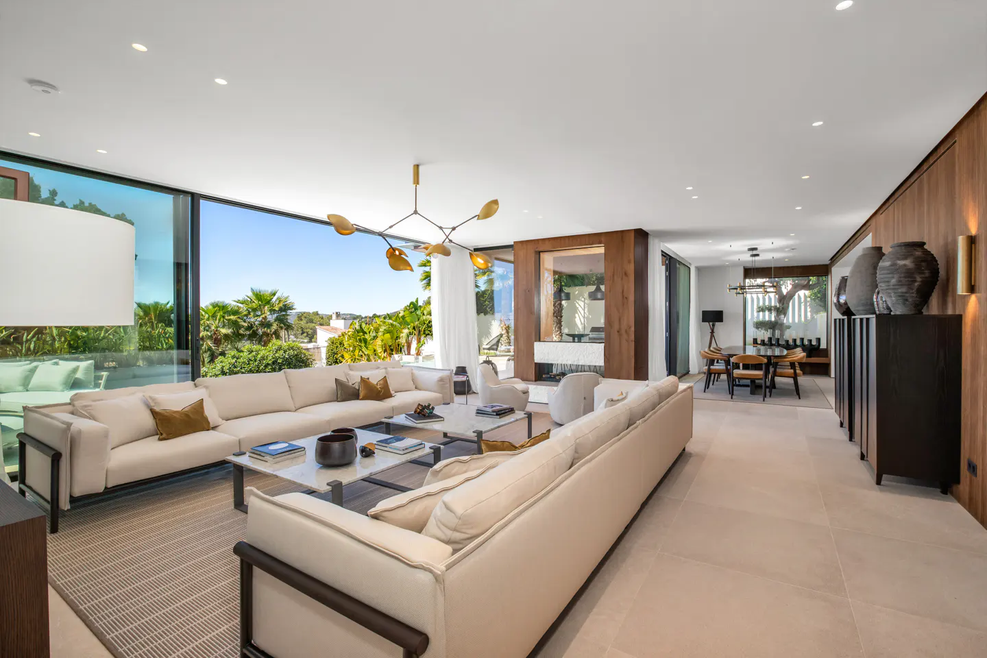 Bright, modern living room with white sofas, marble tables, and a gold chandelier. Large windows offer a view of lush greenery.