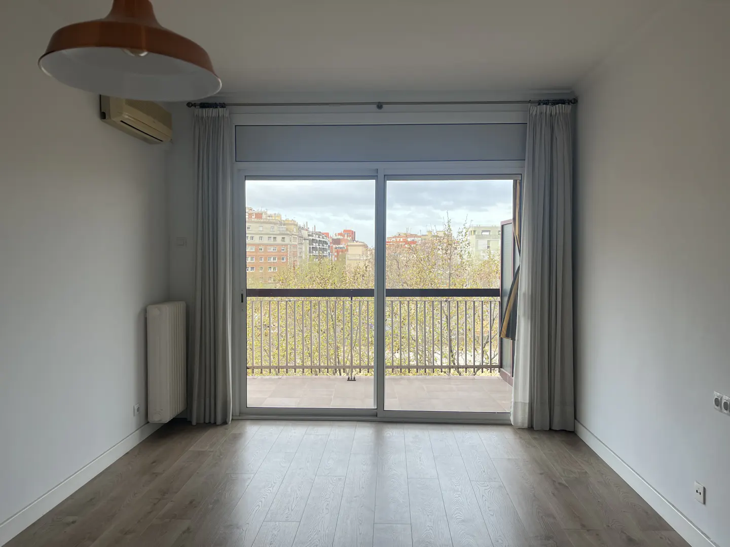 Empty room with light wood floors, white walls, a hanging lamp, and a balcony door overlooking city buildings and trees.