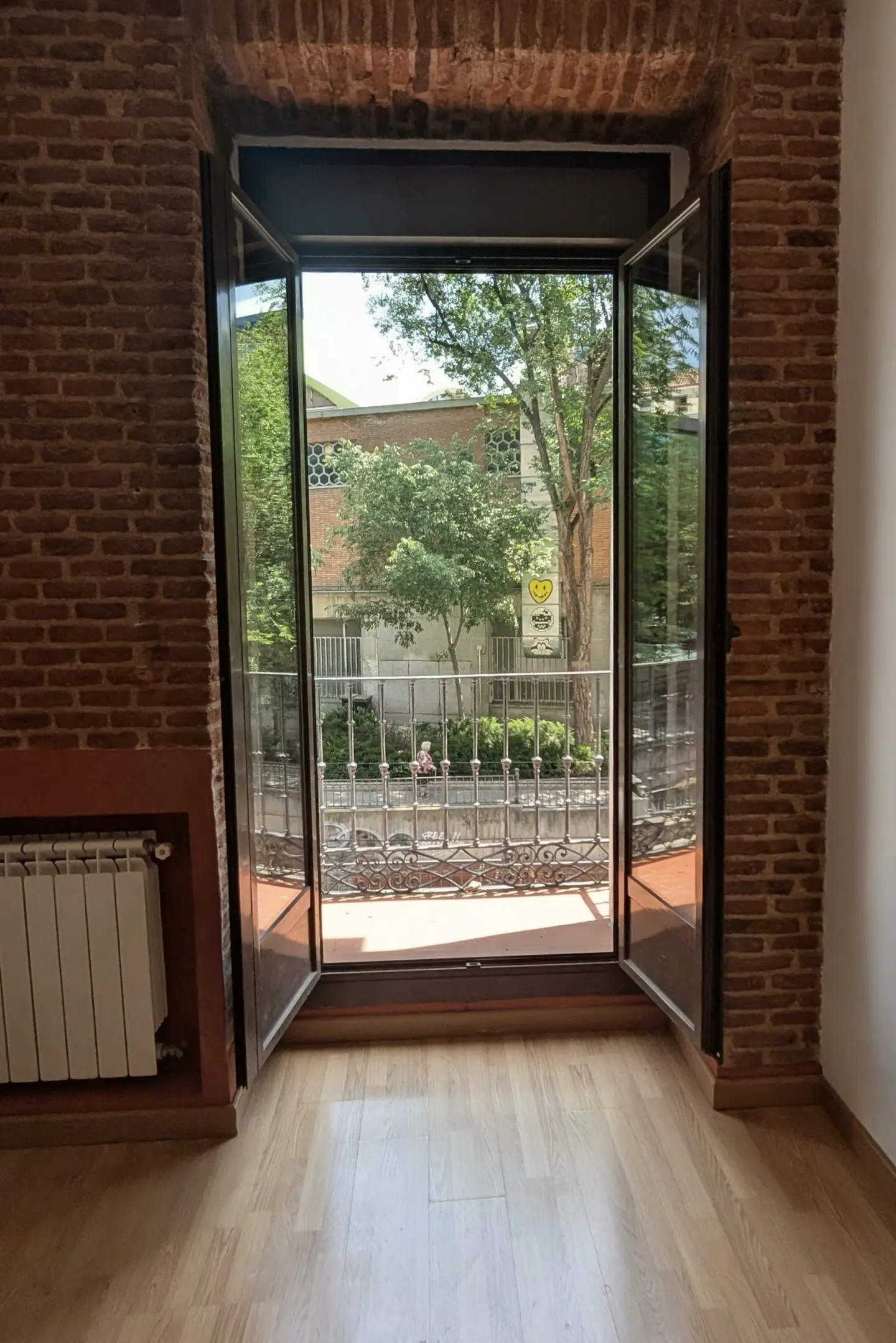 Interior view of a room with exposed brick walls and open doors leading to a balcony with trees and a building in the background.