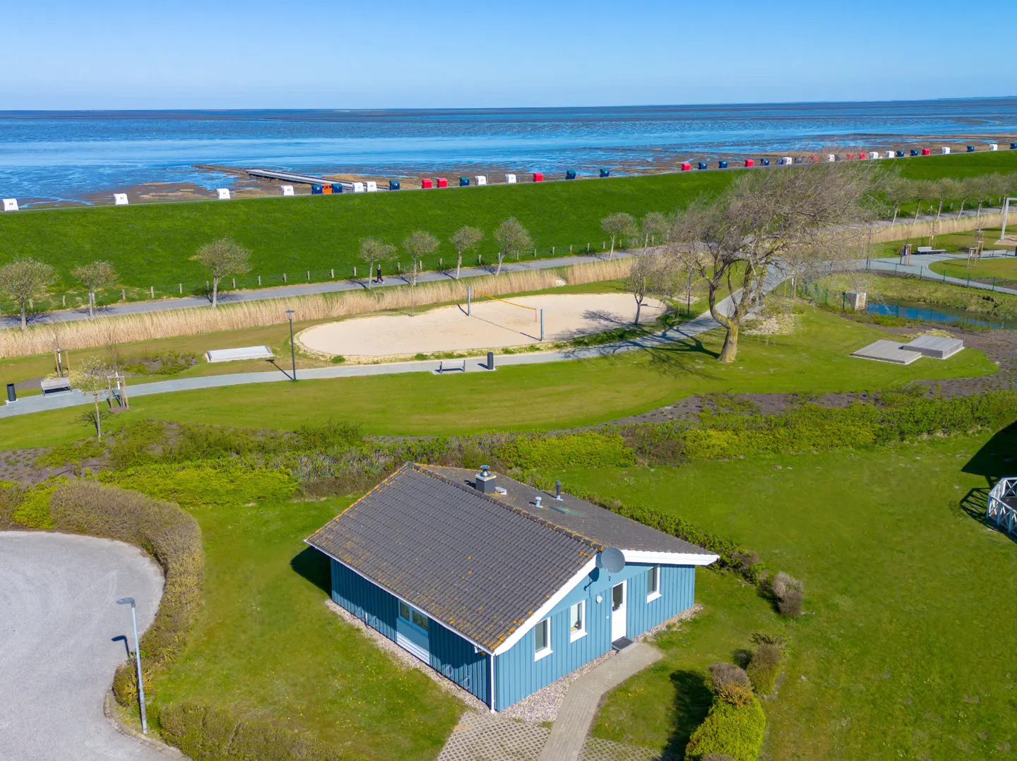 Aerial view of a blue house with a gray roof, green lawn, and a beach with colorful chairs in the background.