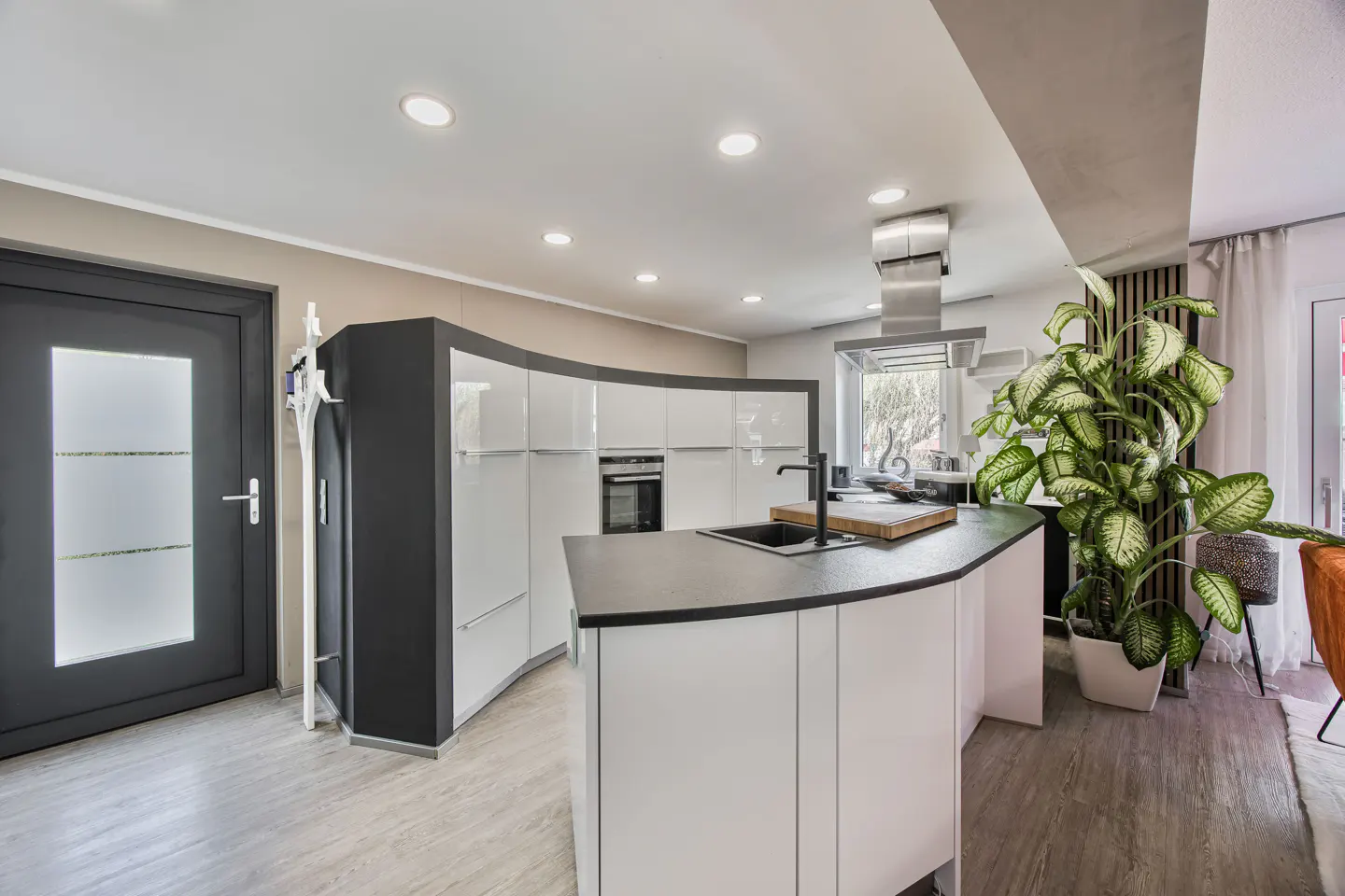 Modern kitchen with curved white cabinets, black countertops, and a stainless steel range hood. A large potted plant adds a touch of nature.