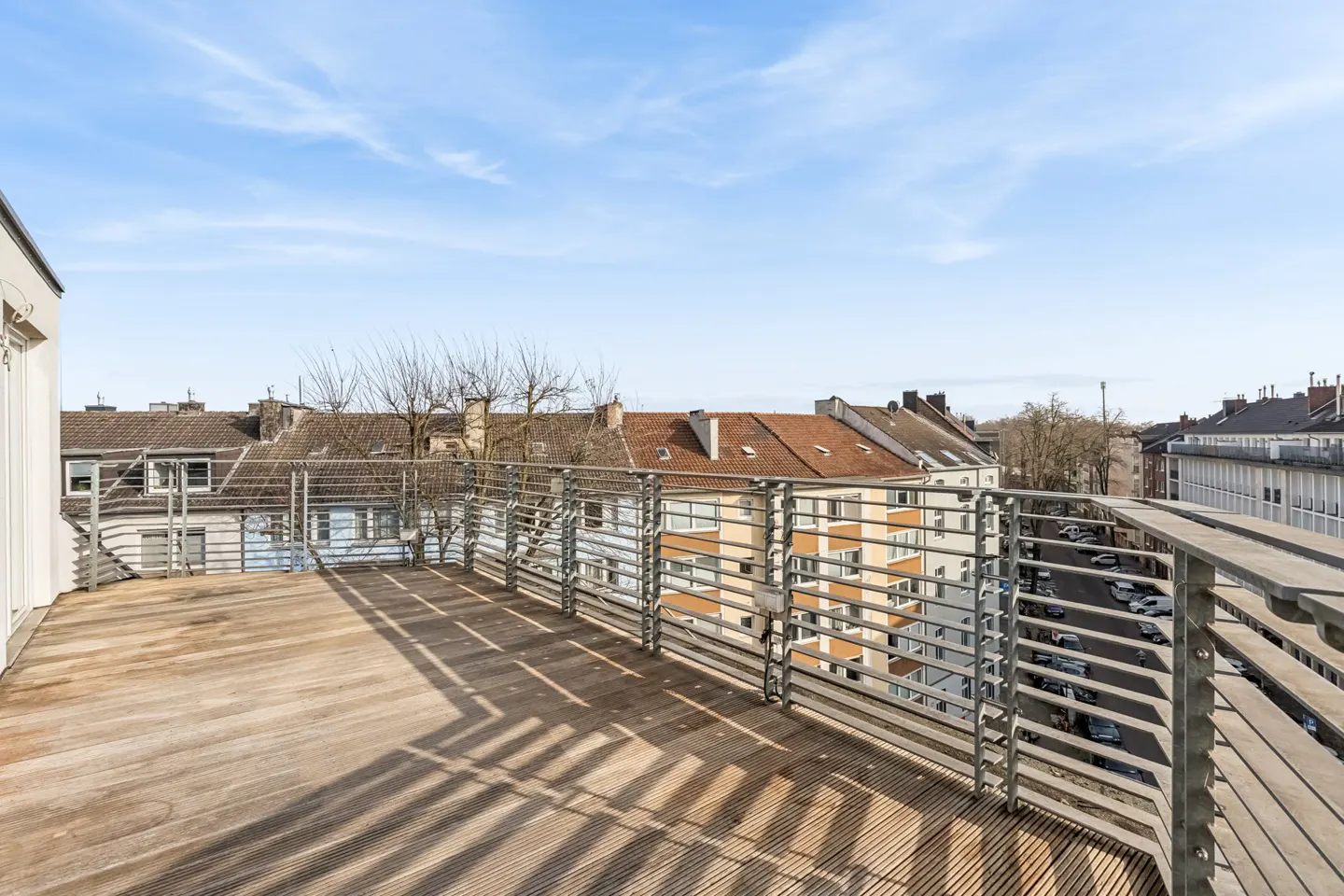 Wide, sunlit wooden deck with metal railings overlooking a European city street and buildings under a blue sky.
