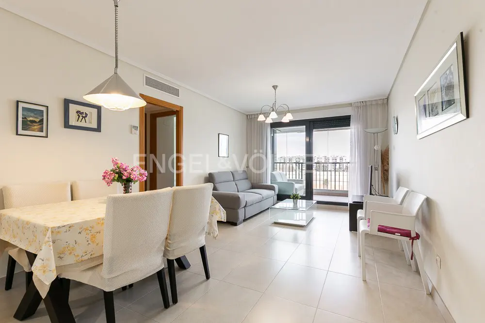Bright living and dining room with a floral tablecloth, grey sofa, glass coffee table, and balcony access.