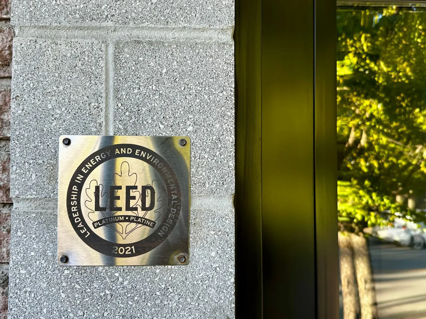 LEED Platinum certification plaque on a gray concrete wall next to a black-framed window with a tree visible through the glass.