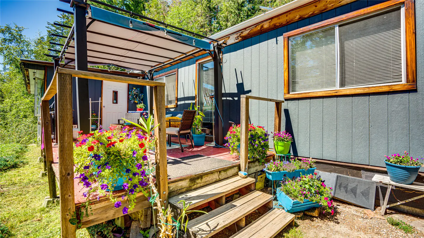 Exterior view of a gray house with a wooden deck, pergola, and colorful flower pots.