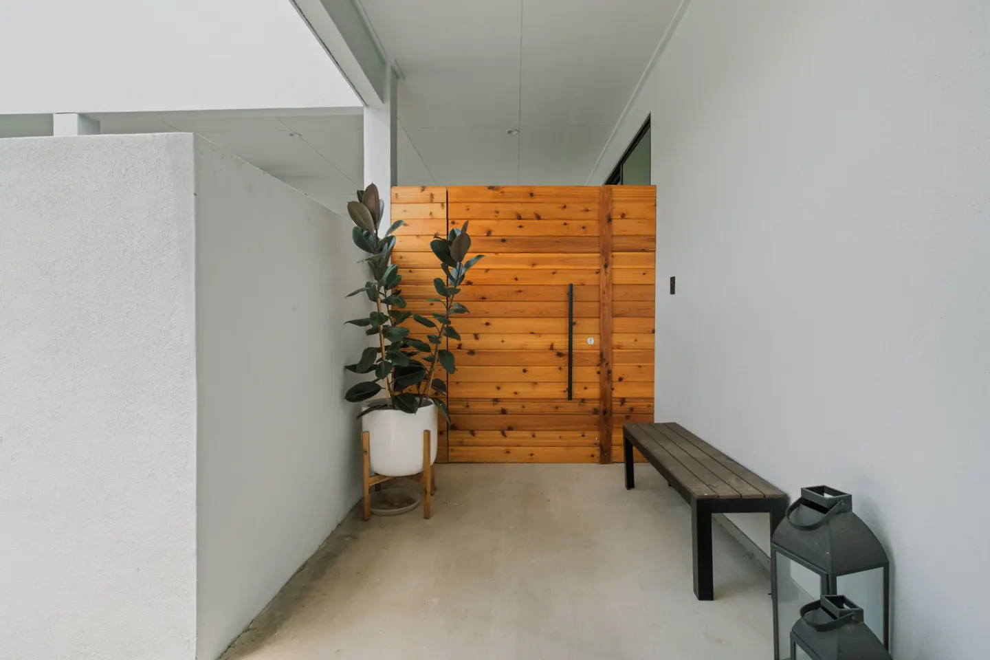 Modern entryway with a horizontal wood door, a potted rubber plant, a wooden bench, and two black lanterns on concrete.