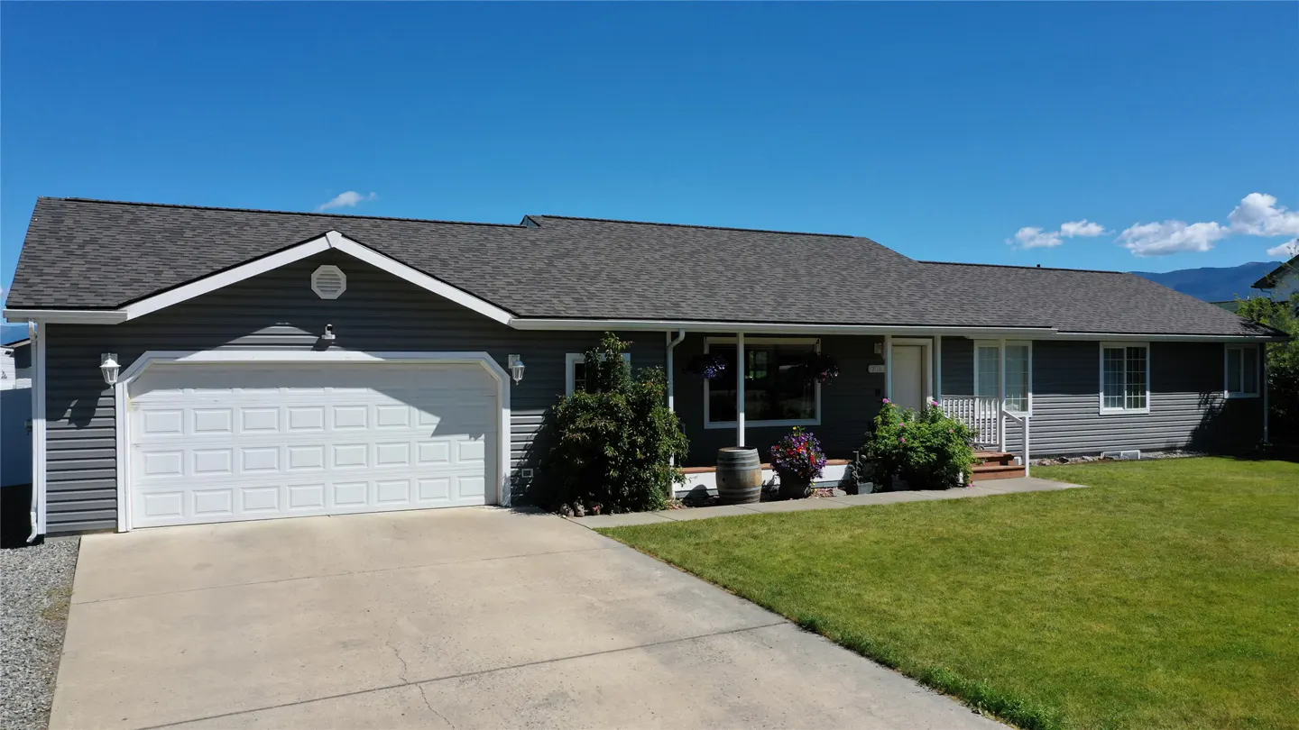 A gray single-story house with a white garage door, green lawn, and blue sky.