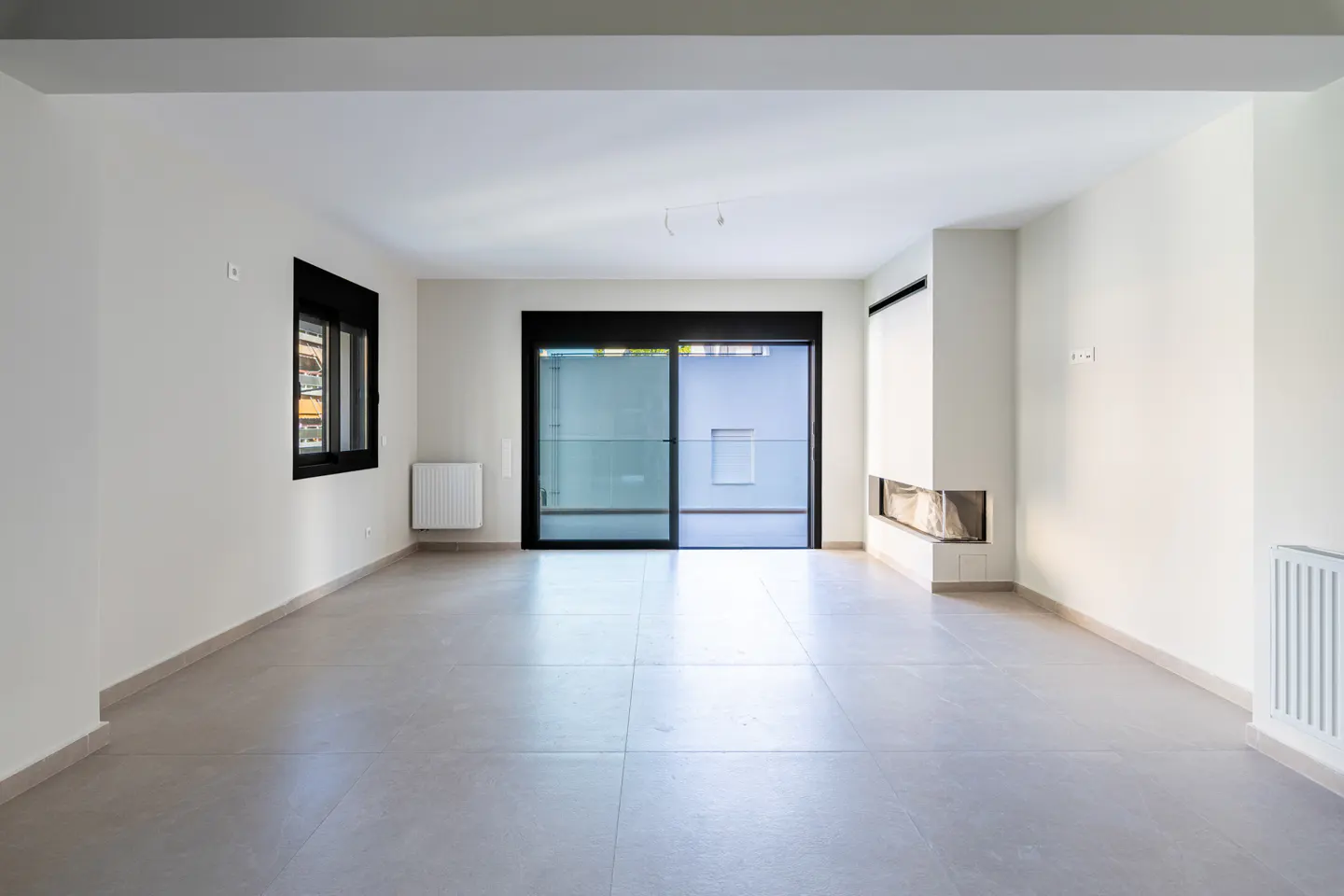 Bright, empty room with white walls, gray tile floor, black-framed window and sliding glass door, and a modern fireplace.