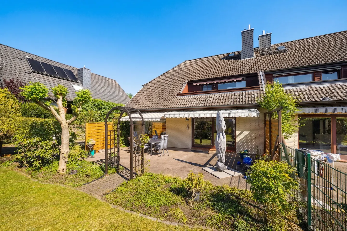 Backyard patio with table and chairs, accessed through an arched trellis. The house has a brown tile roof and a retractable awning.