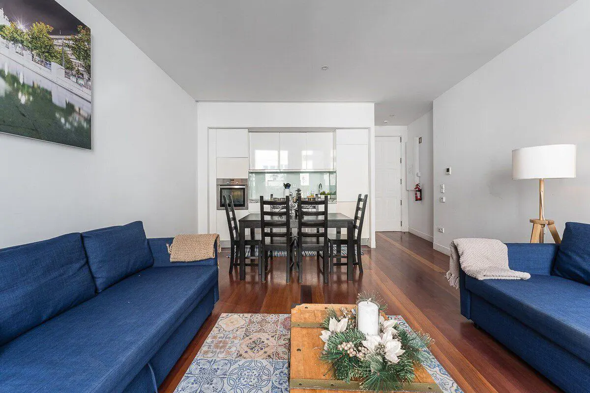 Bright living room with blue sofas, wood floors, and a black dining table. A white kitchen is visible in the background.