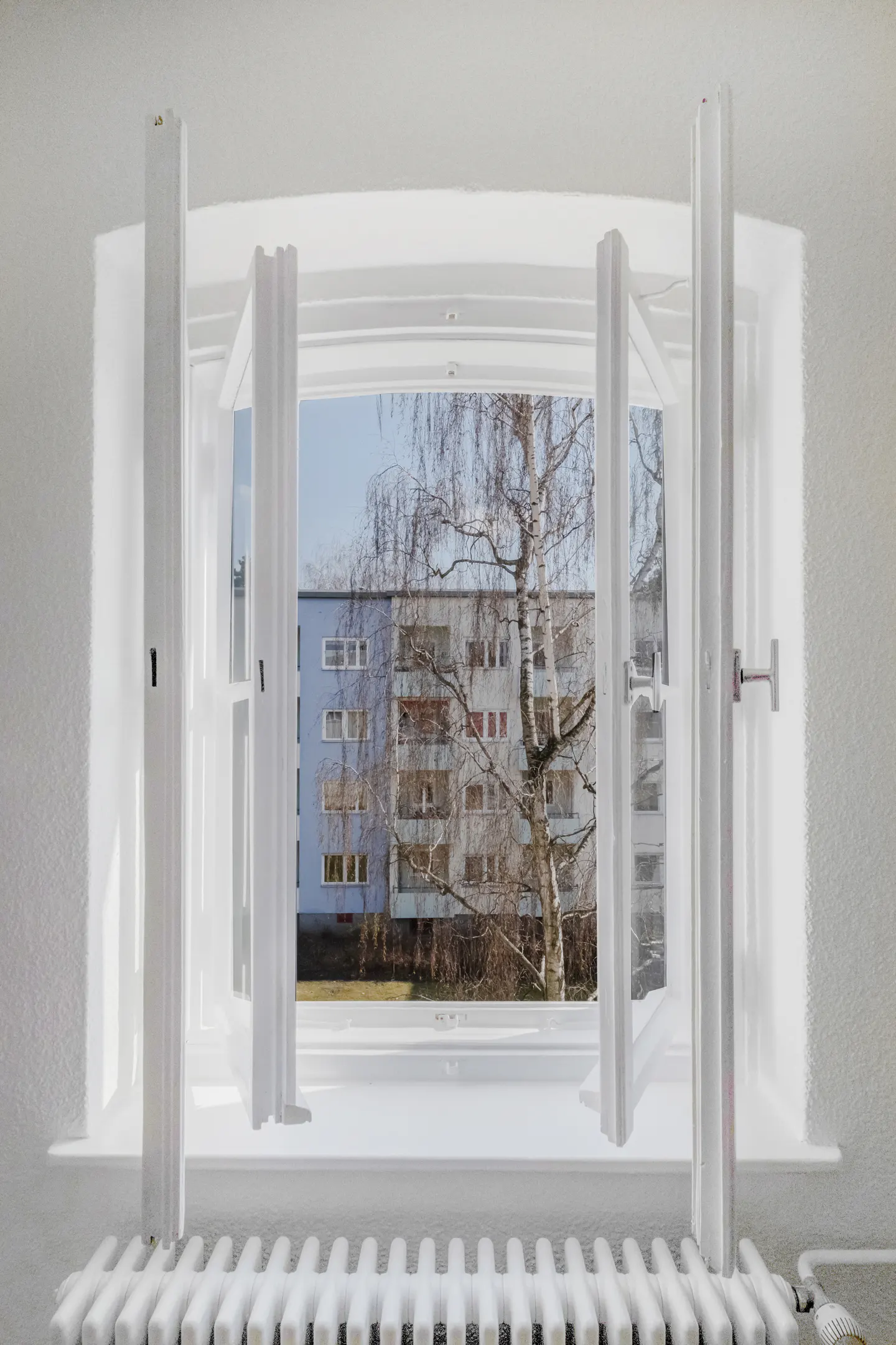 Open white window with a view of a blue apartment building and a tree. A white radiator is below the window.