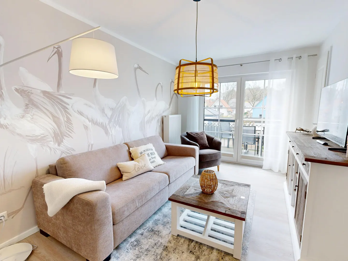Living room with a beige sofa, patterned rug, and a coffee table. A wall mural of cranes adds a unique touch. Balcony access provides natural light.