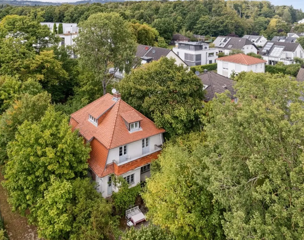 Aerial view of a two-story white house with a red tile roof, surrounded by lush green trees.