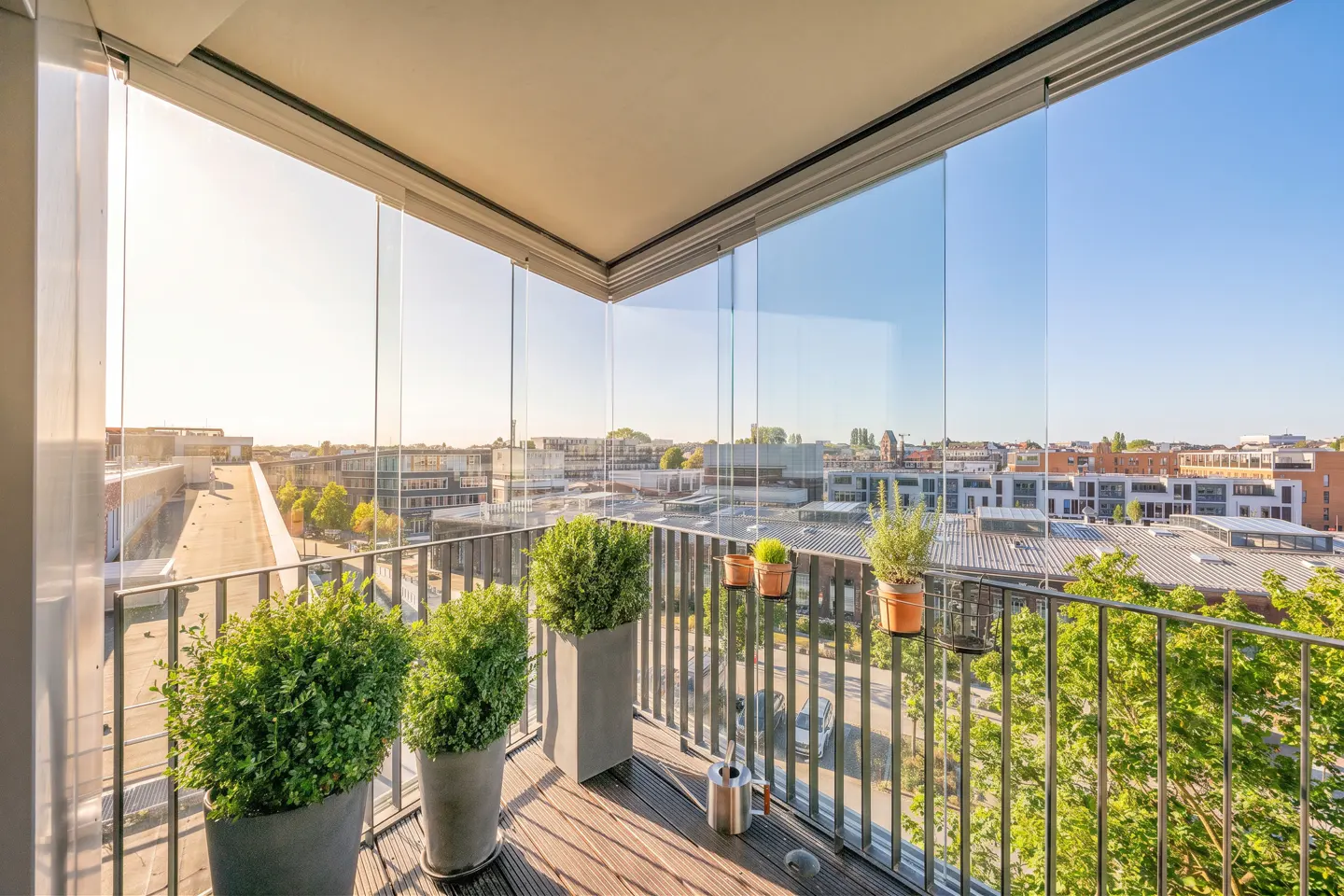 Modern balcony with glass railings, potted plants, and a metal watering can overlooking a sunny urban neighborhood.