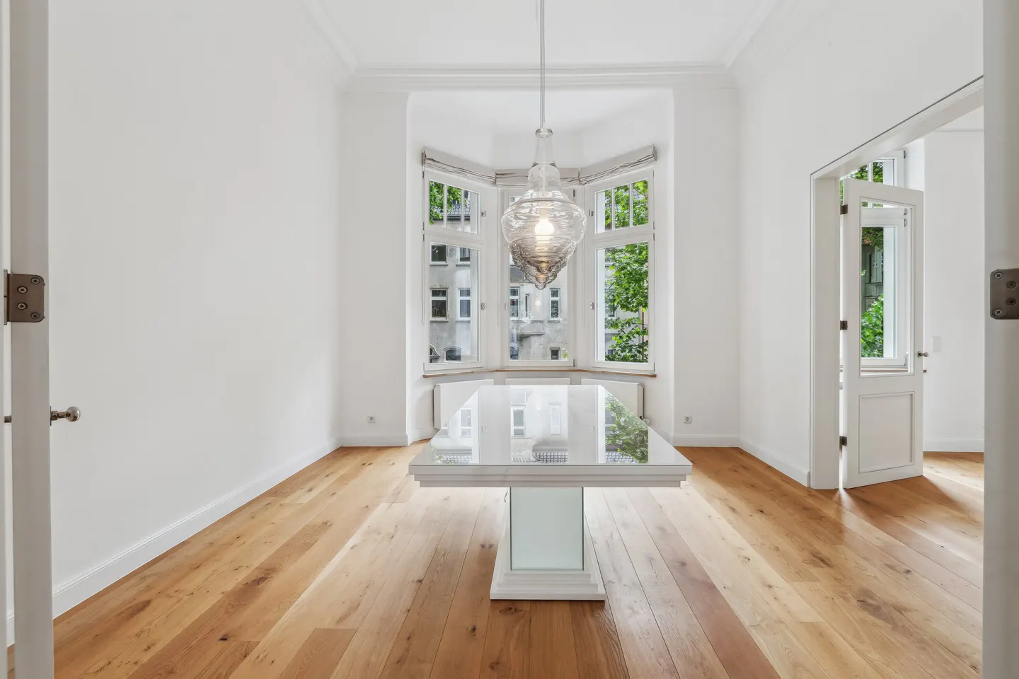 Bright, empty dining room with wood floors, white walls, and a glass table. A modern chandelier hangs above the table, and a bay window offers a view of trees.