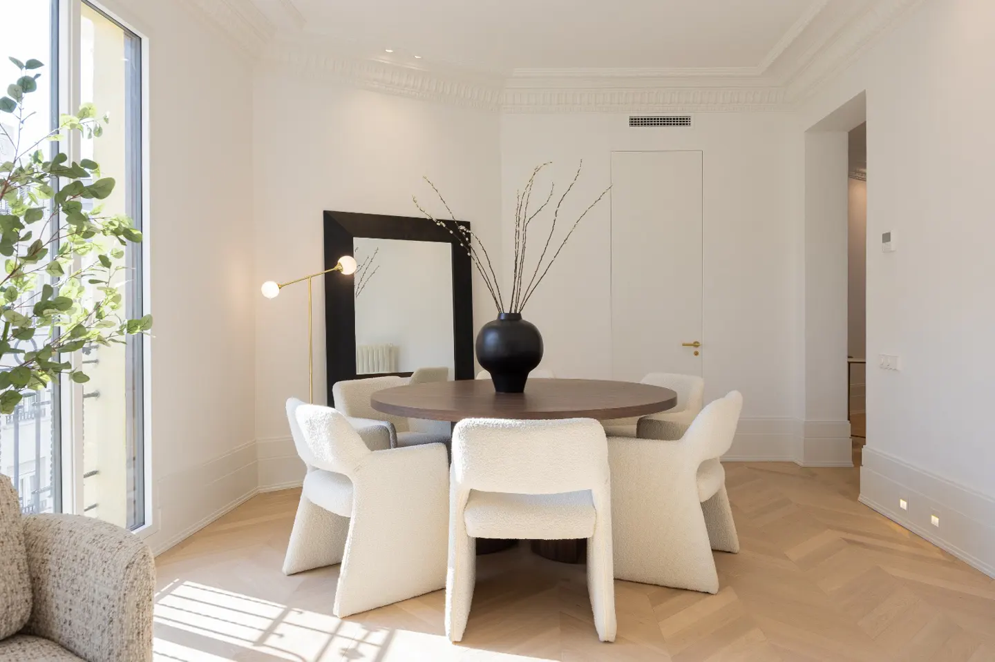 Bright dining room with a round wood table, six white chairs, black vase, and a black framed mirror.