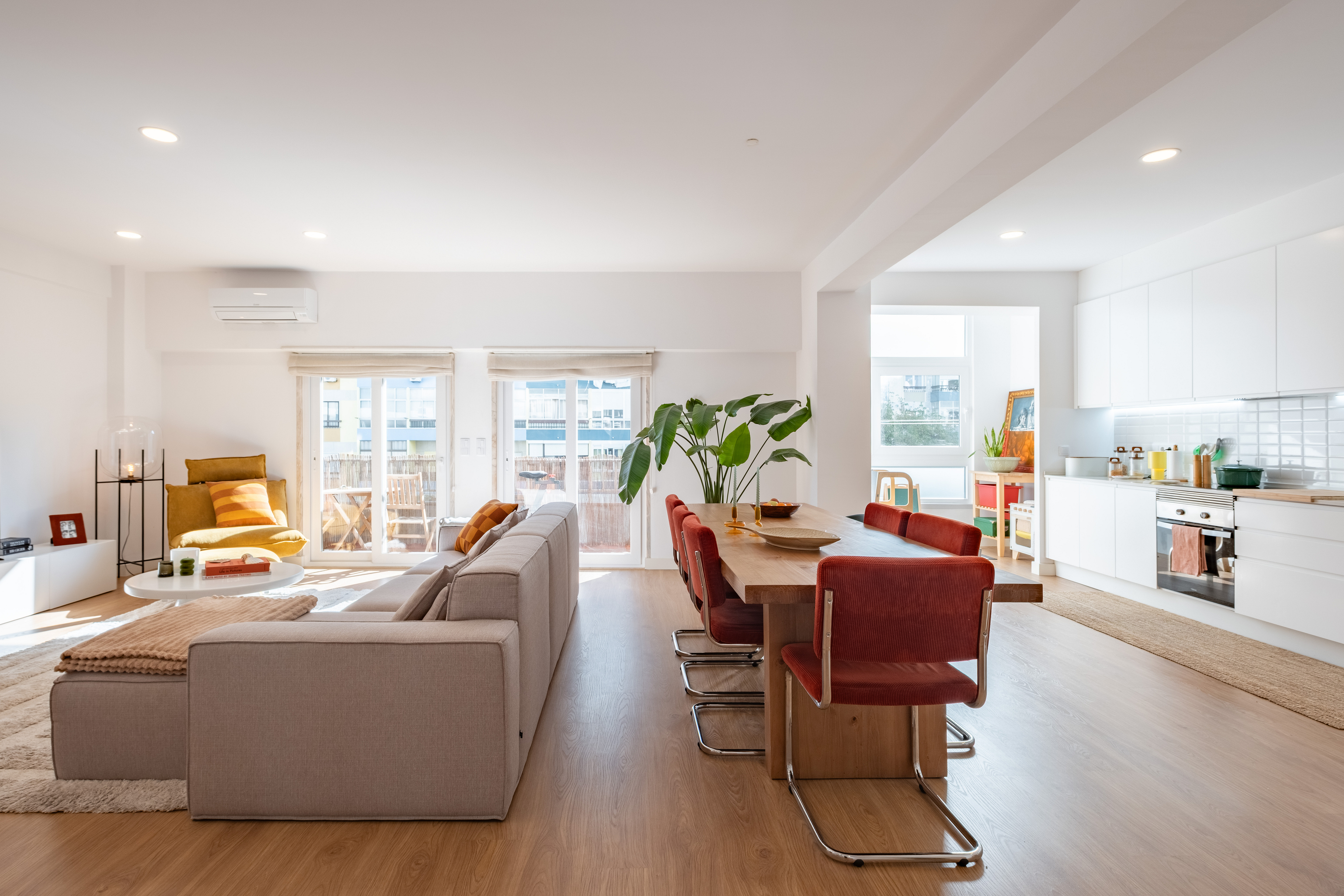 Open-plan living area with a grey sofa, wooden dining table with red chairs, and a modern white kitchen. [AI-generated text]