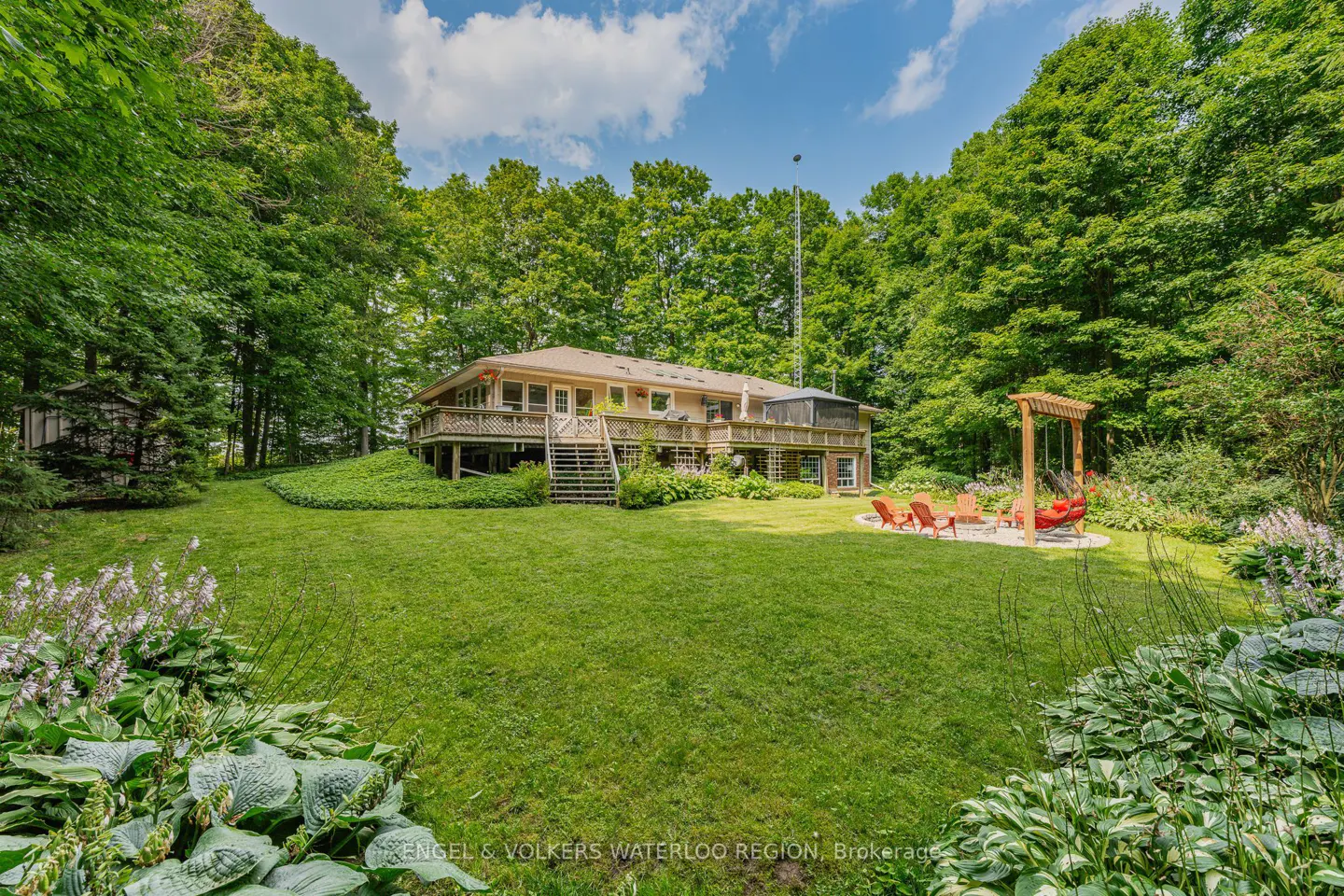 A tan house with a deck overlooks a green lawn, surrounded by trees. A swing set and chairs sit on a gravel area.