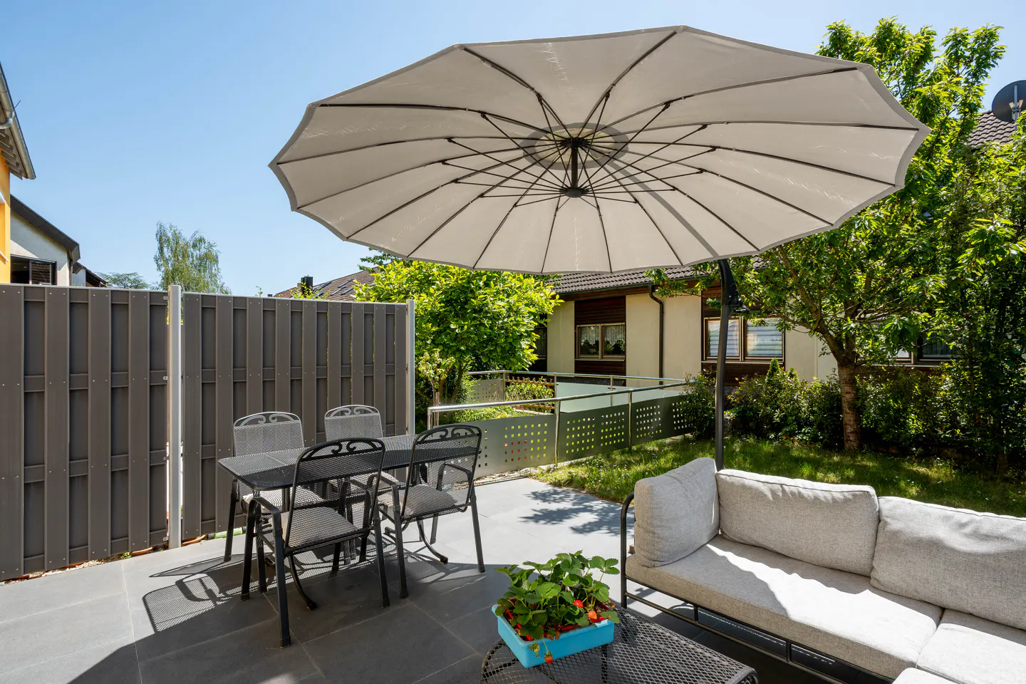 Sunny patio with a large beige umbrella, dining set, and comfortable sofa on a paved terrace next to a garden.