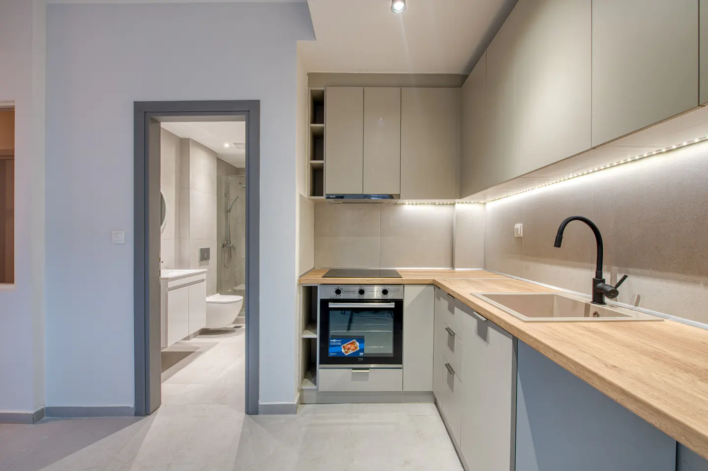 A modern kitchen with gray cabinets, wood countertops, and a black faucet. A bathroom is visible through a doorway.