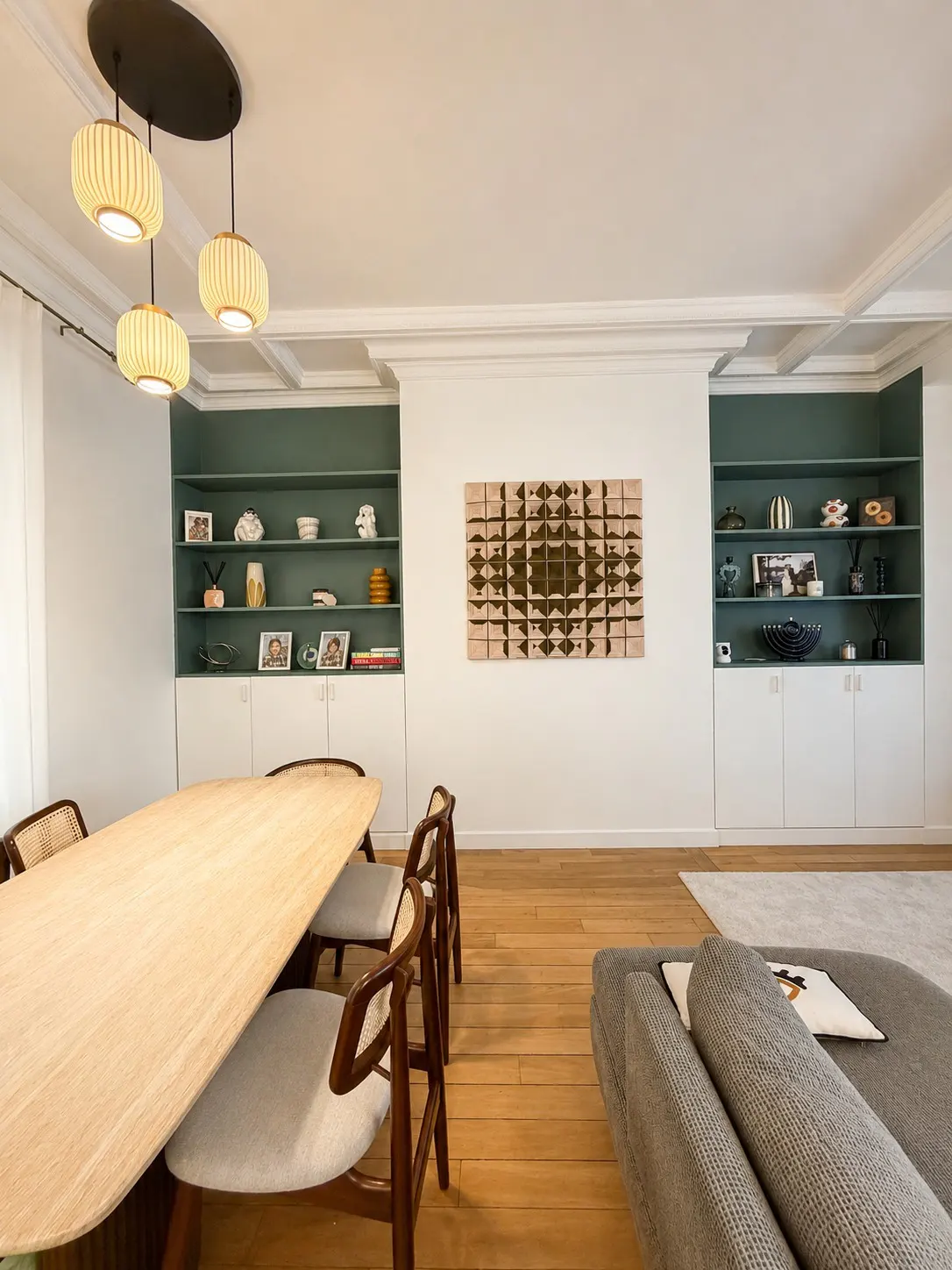 A bright dining room with a light wood table, chairs, and hardwood floors. Built-in shelves with green backing flank a geometric wall art piece.