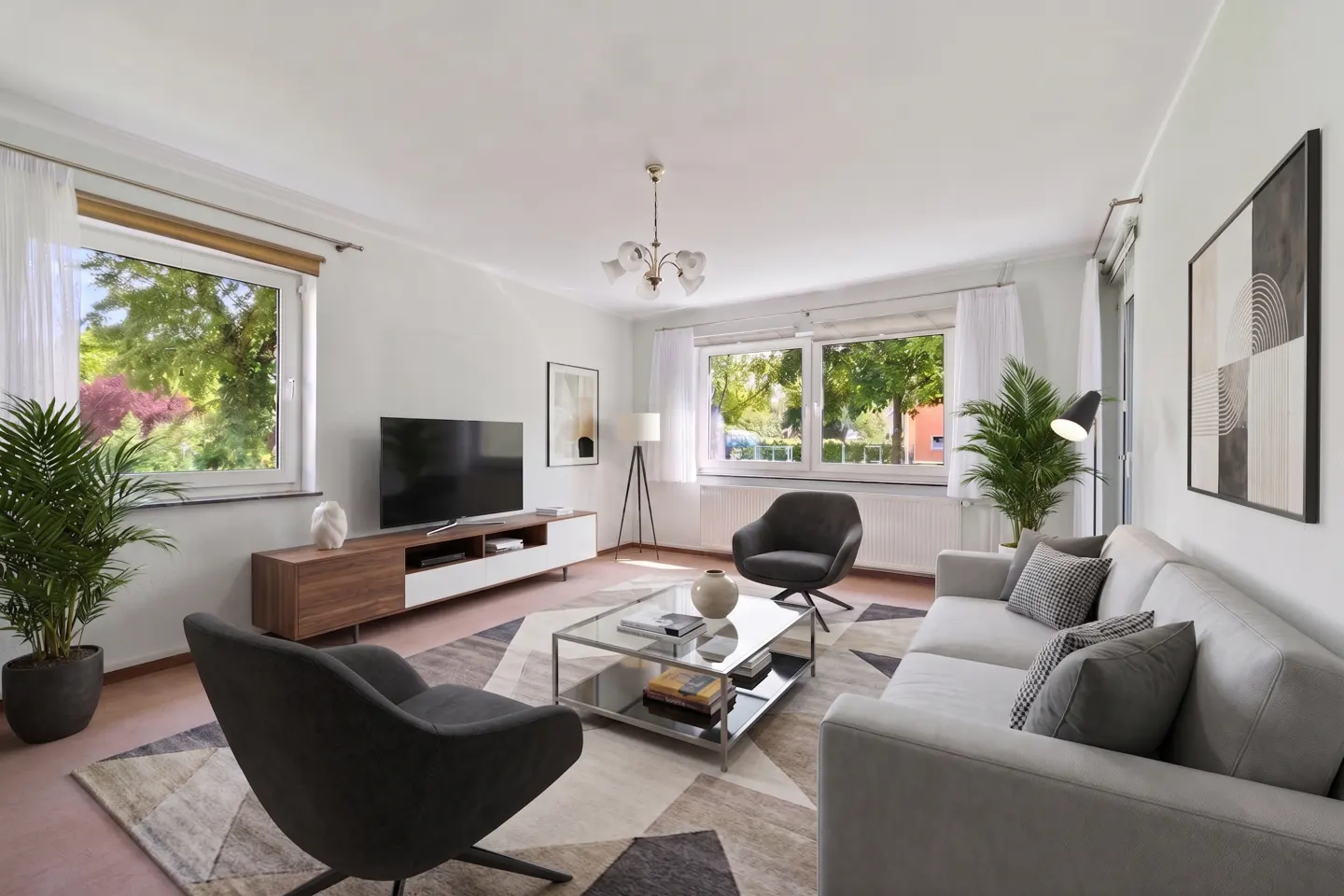 Living room with gray sofa, chairs, and geometric rug. Windows show green trees. TV on a wood and white stand. Modern chandelier.