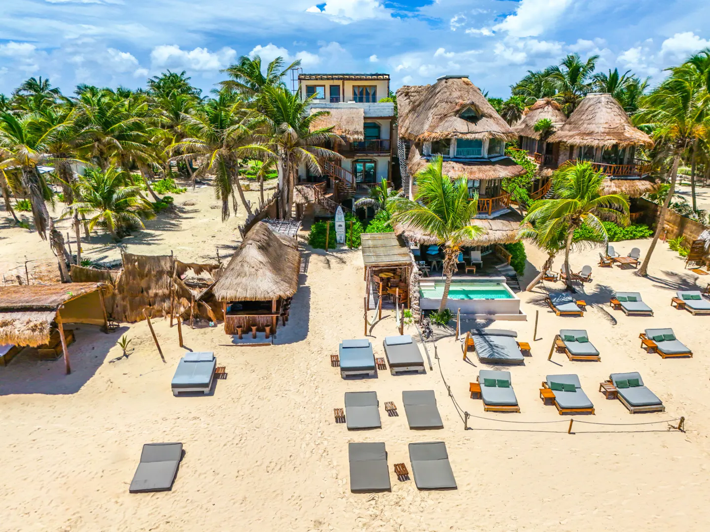Beachfront property with thatched-roof buildings, palm trees, and lounge chairs on white sand under a blue sky.