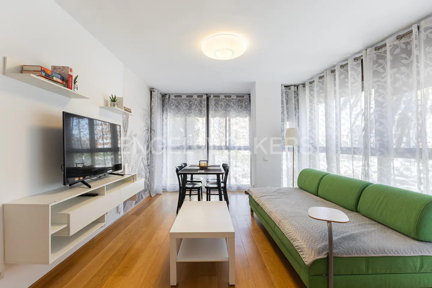 Bright living room with wood floors, green sofa, white shelves, TV, and dining table near sheer curtains.