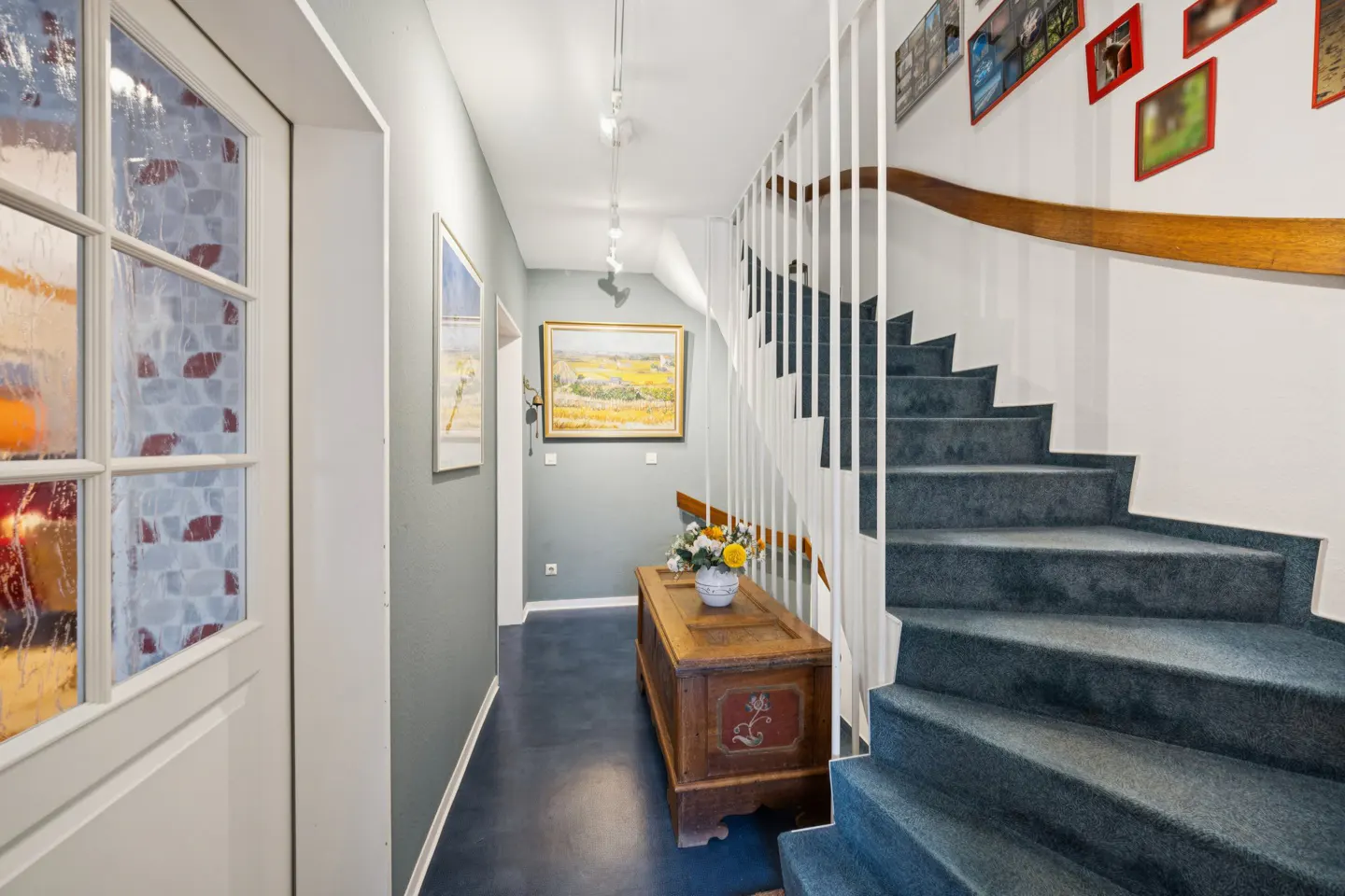 Hallway with blue carpeted stairs, white railings, and a wooden chest with flowers. Paintings adorn the gray walls.