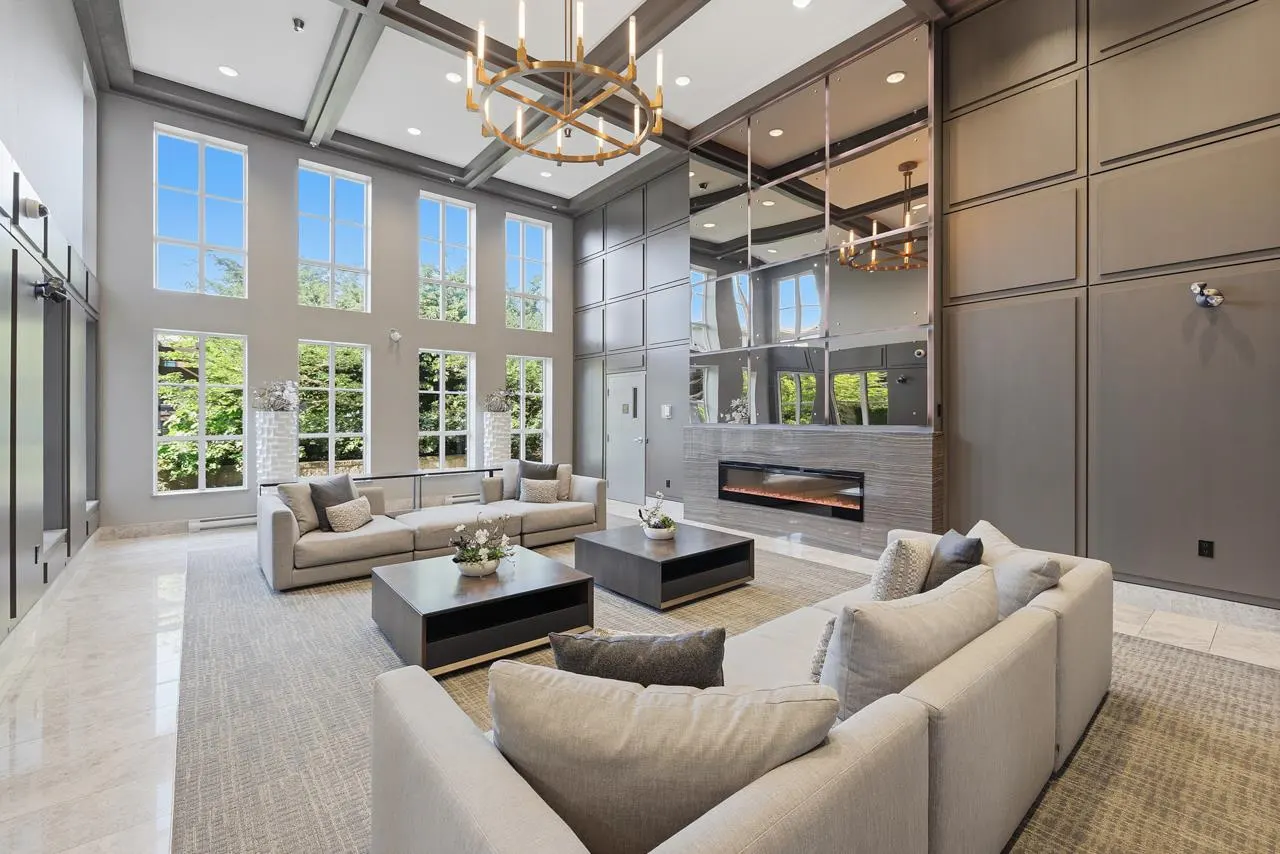 A modern gray lobby with high ceilings, large windows, and neutral-toned sofas. A fireplace and a mirrored wall add elegance.