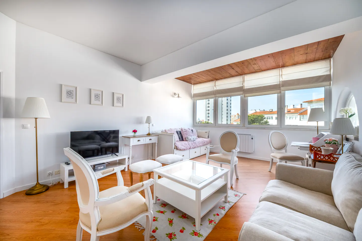Bright living room with white walls, wood floors, and large windows. White furniture includes a sofa, chairs, and a coffee table with a floral rug.