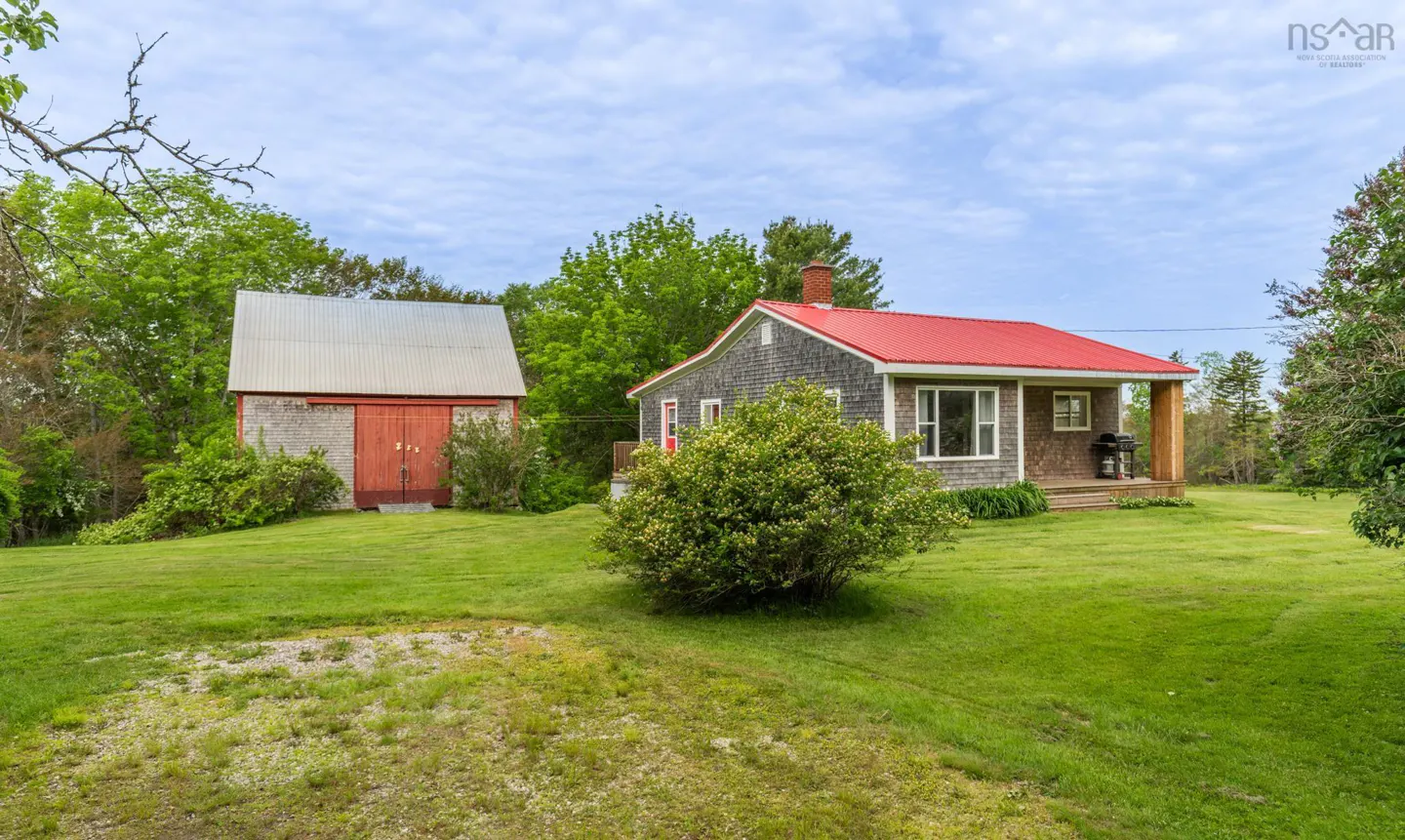 A grey shingled house with a bright red roof and a matching detached barn sit on a large, grassy lot under a blue sky.
