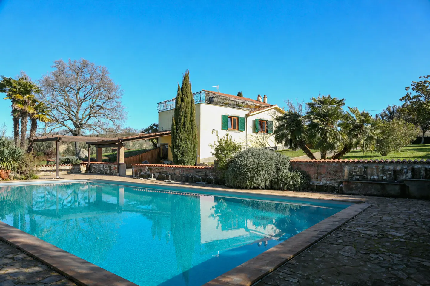 A sunny view of a turquoise pool in front of a cream-colored house with green shutters and a stone wall.