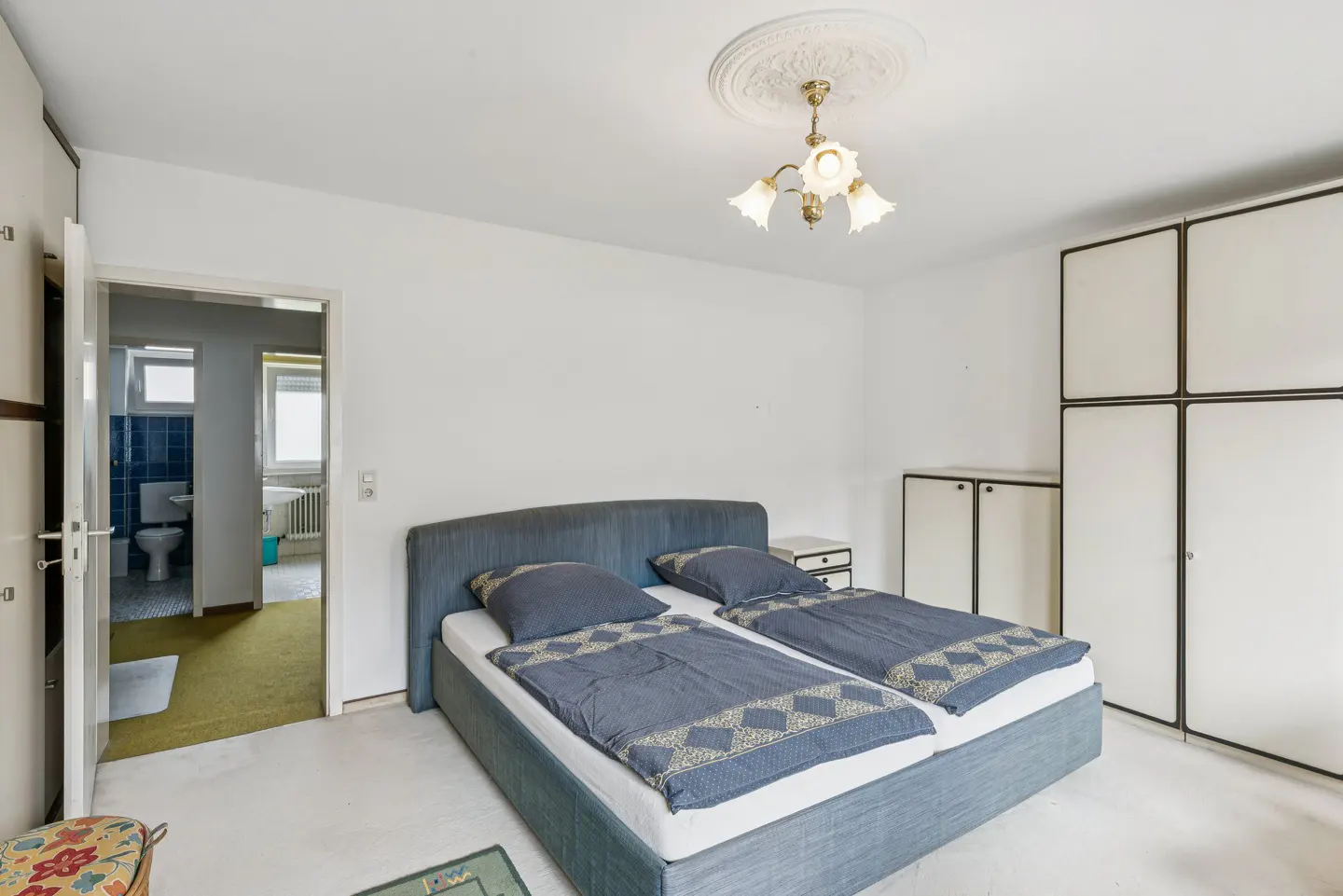Bedroom with a blue bed, white walls, and a view into the bathroom. A chandelier hangs from the ceiling.