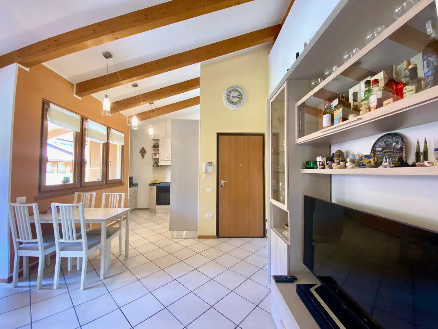 Interior view of a living and dining area with white tile floors, wooden ceiling beams, and a dining table with white chairs. A kitchen is visible in the background.