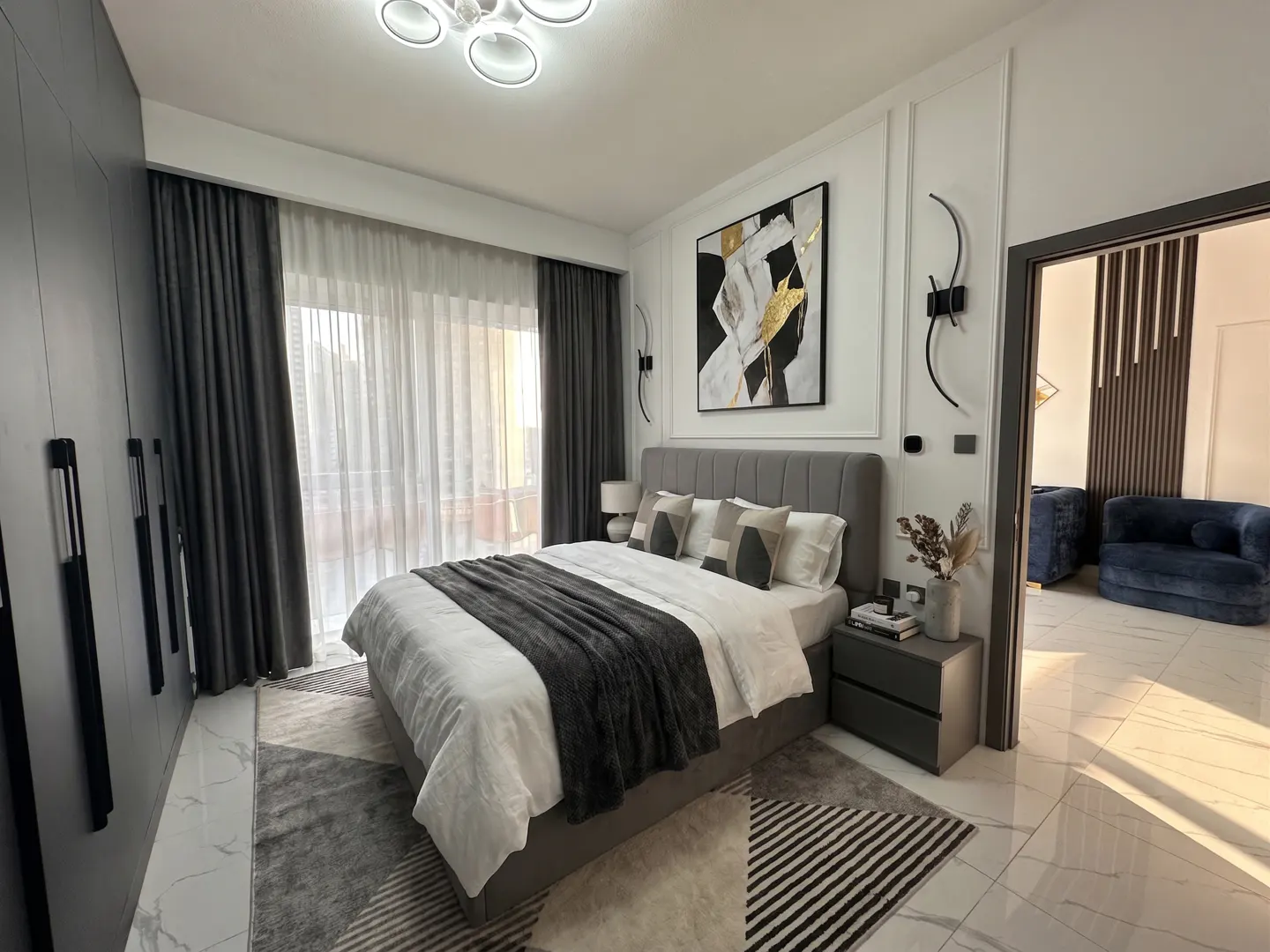 Modern bedroom with a grey upholstered bed, white bedding, dark curtains, geometric rug, and abstract wall art.