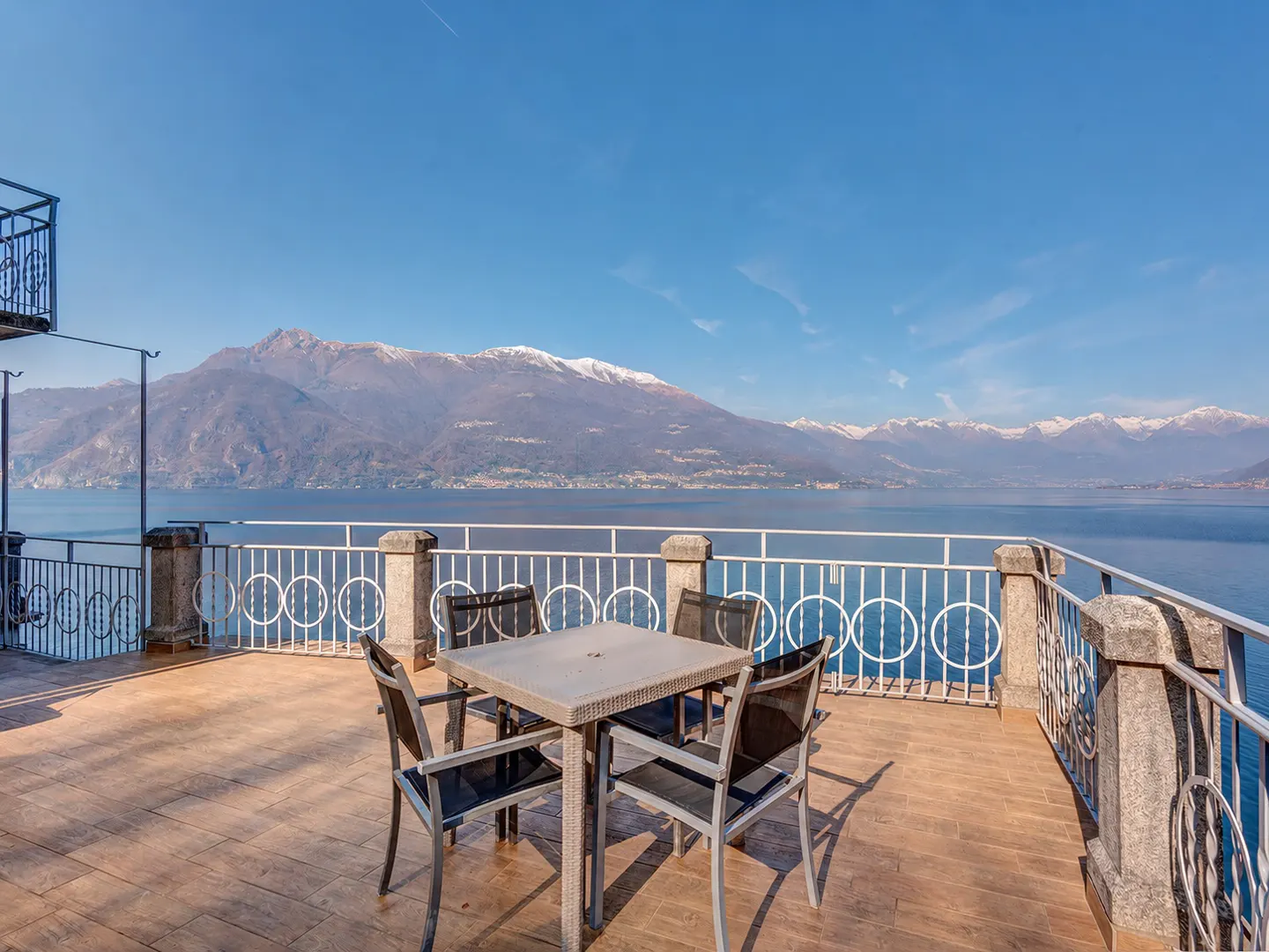 Outdoor patio with table and chairs overlooking a lake and mountains under a blue sky.
