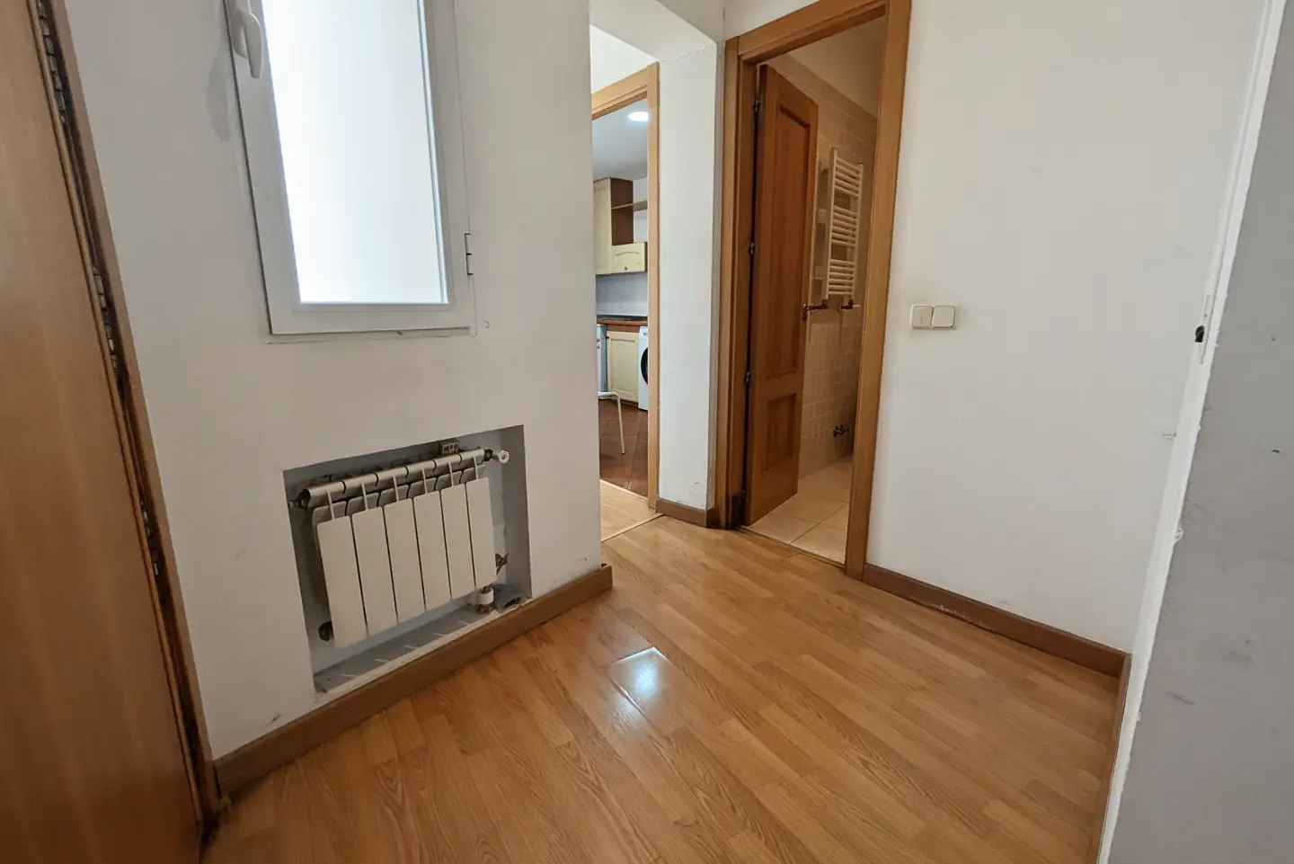 Hallway with wood floors, white walls, and two open doorways. One leads to a kitchen, the other to a bathroom. A radiator sits under a window.