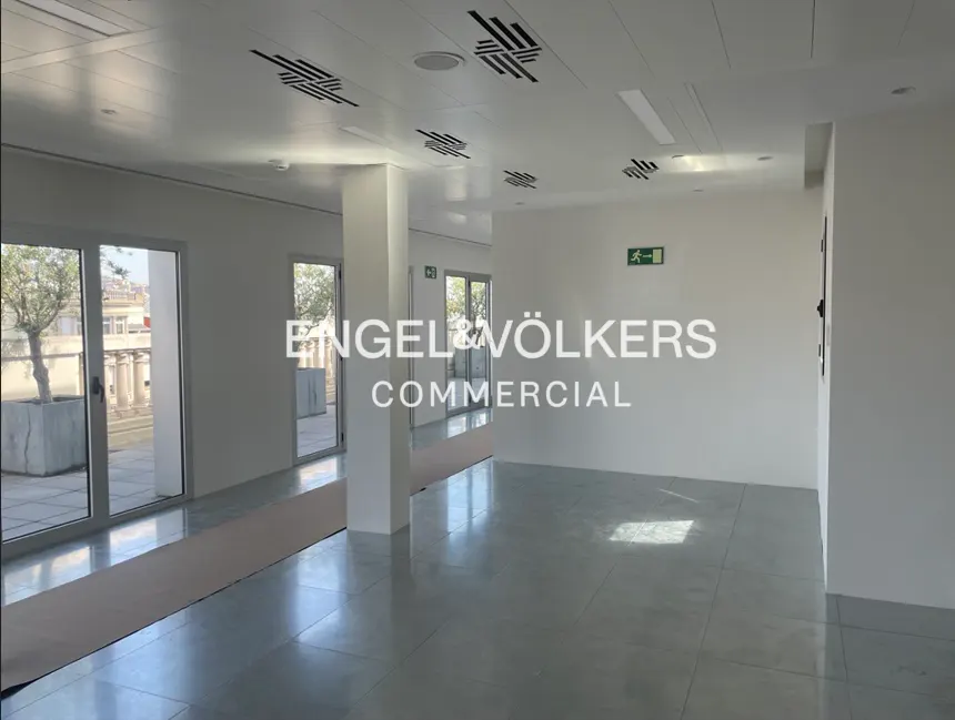 Bright, empty commercial space with white walls, gray floors, and large windows. Engel & Völkers Commercial logo on the wall.